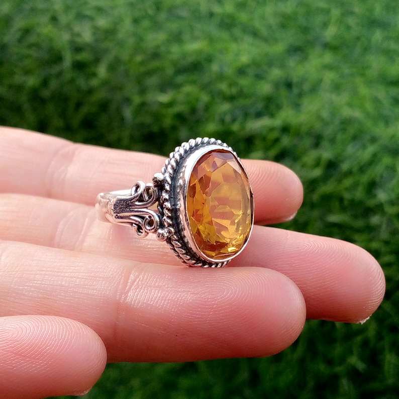 May include: A silver ring with an oval, amber-colored gemstone. The gemstone is set in a decorative bezel with a twisted rope design. The ring's band features ornate scrollwork on the sides, creating a vintage aesthetic.