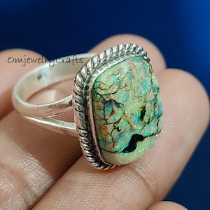 May include: A silver ring with a large, rectangular opal gemstone. The opal has a unique pattern of green, blue, and black colors. The ring band is simple, while the gemstone is framed by a textured silver border. The text "Omjewelrycrafts" is visible.