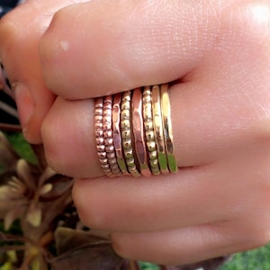 May include: A selection of gold and copper rings stacked on a finger. The rings have different textures, including beaded, hammered, and smooth finishes. The rings are shown on a hand with a blurred background.