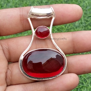 Op de afbeelding: Zilveren hanger met twee rode edelstenen: een groot ovaal aan de onderkant en een kleinere, ronde erboven. De hanger heeft een kenmerkend, open ontwerp met een lus aan de bovenkant. De achtergrond is een wazig groen.