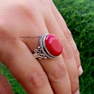 以下が含まれることがあります： 大きな楕円形の鮮やかな赤い宝石が付いたシルバーリング。リングは、装飾的な渦巻きと編み込みのボーダーが付いた、装飾的で質感のあるシルバーバンドです。リングは人の指にあります。