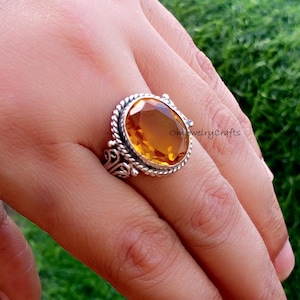 Può includere: Un anello d'argento con una gemma ovale color ambra. L'anello ha un bordo a forma di corda attorno alla pietra e filigrane decorative sui lati. L'anello è sul dito di una persona.