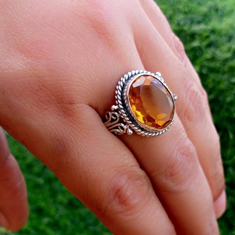May include: A silver ring featuring a large, oval-cut, amber-colored gemstone. The ring has an ornate, detailed design with a twisted rope border around the stone. The ring is set on a hand, showcasing the jewelry.