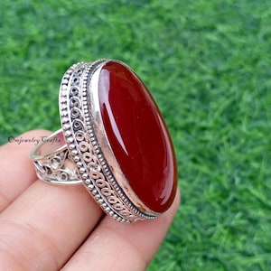 Könnte beinhalten: Silberring mit einem ovalen, polierten, dunkelroten Edelstein. Der Ring hat ein verziertes, detailliertes Design um den Edelstein und am Band. Der Text "Omjewelry Crafts" ist sichtbar.