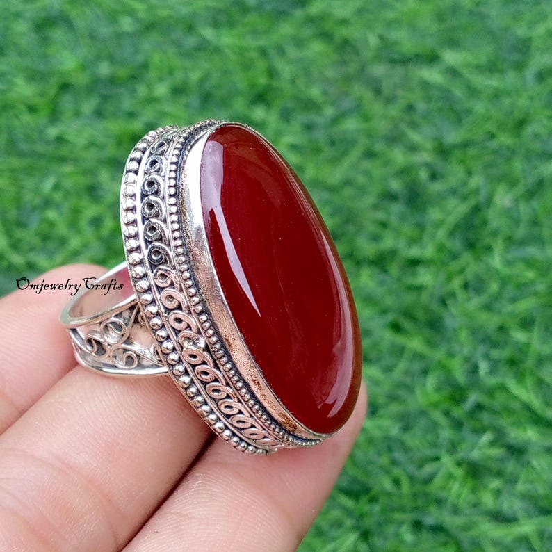 May include: A silver ring with an oval, polished, dark red gemstone. The ring has an ornate, detailed design around the gemstone and on the band. The text "Omjewelry Crafts" is visible on the ring.
