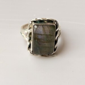 Op de afbeelding: Zilveren ring met een rechthoekige, iriserende, grijsgroene steen. De steen is gezet in een gevlochten, touwachtige rand. De ring heeft een eenvoudige band en wordt gepresenteerd op een wit oppervlak.