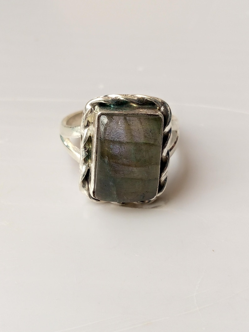 Op de afbeelding: Zilveren ring met een rechthoekige, iriserende, grijsgroene steen. De steen is gezet in een gevlochten, touwachtige rand. De ring heeft een eenvoudige band en wordt gepresenteerd op een wit oppervlak.