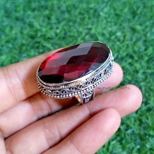 Op de afbeelding: Een sierlijke ring met een grote, ovale, dieprode edelsteen. De zilverkleurige band heeft gedetailleerde patronen. De ring wordt in een hand gehouden tegen een groene achtergrond. De tekst "omjewelrycrafts" is zichtbaar op de ring.