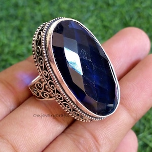 May include: A large, oval-shaped ring with a faceted, deep blue gemstone. The ring has an ornate, silver-colored band with scrollwork detailing around the gemstone and on the sides. The ring is set against a green background.