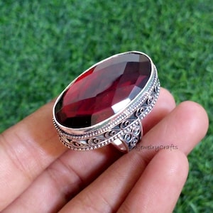 May include: A large, oval-shaped ring with a faceted, deep red gemstone. The ring has an ornate, silver-colored band with detailed scrollwork. The ring is held against a green background.