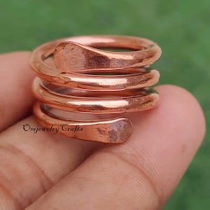 Könnte beinhalten: Ein kupferfarbener Spiralring mit gehämmerten Texturen. Der Ring wickelt sich in einem Spulendesign um den Finger, wobei die Enden abgeflacht sind. Der Ring ist handgefertigt und trägt den Text "Omjewelry Crafts".