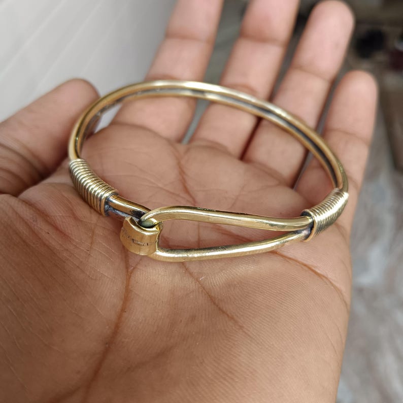 K&ouml;nnte beinhalten: Ein goldfarbenes Armreif-Armband mit einem einzigartigen Verschlussdesign. Das Armband besteht aus zwei Metallteilen, die mit einem verdrehten Draht verbunden sind.