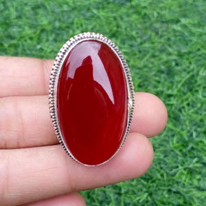 May include: Oval-shaped, red carnelian gemstone ring with a silver-colored, textured border. The polished stone reflects light, set against a green background.