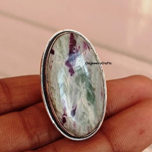May include: Oval-shaped ring with a green, white, and purple gemstone set in a silver band. The ring is on a finger.