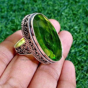 以下が含まれることがあります： 大きな楕円形の緑色の宝石をあしらったシルバーリング。リングは、宝石の周りとバンドに精巧なフィリグリーデザインが施されています。リングは緑色の背景にセットされています。