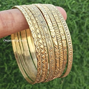 Może przedstawiać: Zestaw ośmiu złotych bransoletek metalowych z misternymi wzorami w reliefie. Bransoletki są ułożone jedna na drugiej i mają różne wzory, w tym kwiatowe, geometryczne i abstrakcyjne.