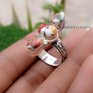 Puede incluir: Un anillo de plata con tres piedras coloridas en forma de lágrima. Las piedras muestran tonos rojos, blancos, naranjas y turquesas. La banda del anillo tiene un diseño floral detallado.