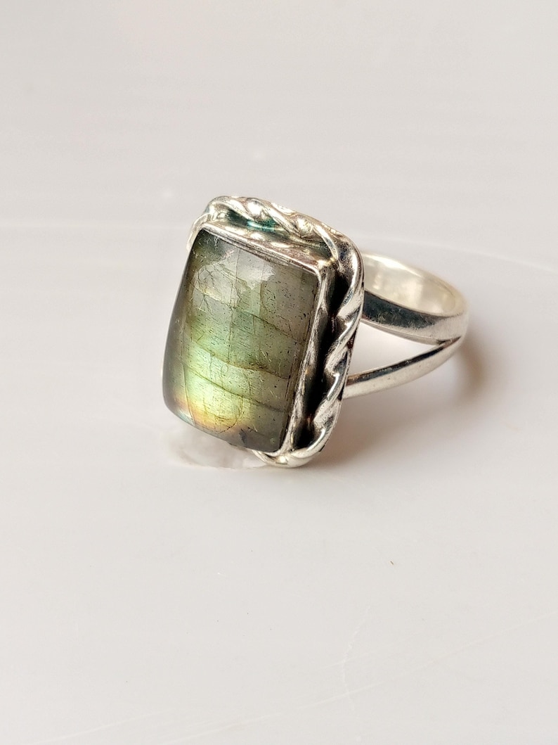 Op de afbeelding: Zilveren ring met een rechthoekige, iriserende labradoriet edelsteen. De steen vertoont tinten groen, geel en blauw. De ring heeft een gevlochten ontwerp rond de steen en een gespleten schacht.