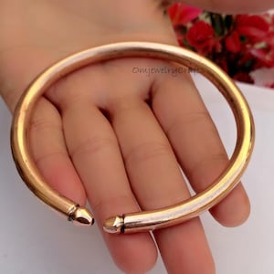 Puede incluir: Un brazalete simple de cobre con extremos redondeados. El brazalete está abierto y tiene un acabado liso y pulido.