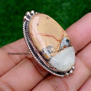 May include: An oval-shaped ring featuring a large, smooth, multi-coloured stone with beige, white, and dark grey inclusions. The stone is set in a polished silver-tone metal frame with a twisted rope border and small decorative silver spheres.