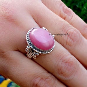 Puede incluir: Anillo de plata con una gran gema rosa ovalada. El anillo tiene una banda de plata decorativa con un borde en forma de cuerda y detalles ornamentados. El anillo se muestra en un dedo. El anillo es una joya.