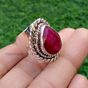May include: A silver ring featuring a large, teardrop-shaped, deep red gemstone. The gemstone is set in a decorative, textured bezel. The ring is held in a hand against a blurred green background. The text "OMJEWELRYCRAFT" is visible.