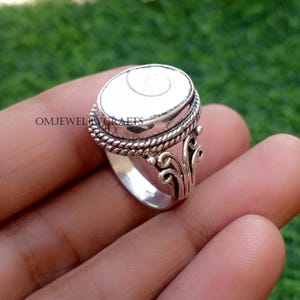May include: A silver ring with an oval, white stone featuring a spiral design. The ring has a braided border around the stone and decorative flourishes on the sides. The text "OMJEWELCRAFTS" is visible on the side of the ring.