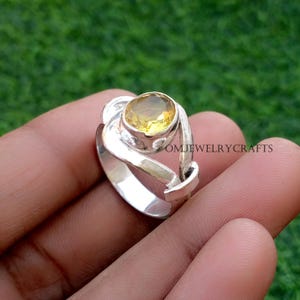 Può includere: Un anello d'argento con una grande pietra preziosa gialla ovale. L'anello ha un design unico e a spirale attorno alla pietra. Il testo "OMJEWELRYCRAFTS" è visibile sul lato dell'anello.