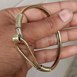 K&ouml;nnte beinhalten: Ein goldfarbenes Armreif-Armband mit einem einzigartigen gedrehten Design. Das Armband besteht aus zwei Metallteilen, die miteinander verdreht sind, um einen einzigartigen Look zu erzeugen. Das Armband ist offen und kann an verschiedene Handgelenksgr&ouml;&szlig;en angepasst werden.
