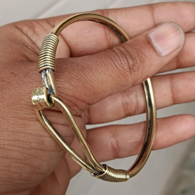 K&ouml;nnte beinhalten: Ein goldfarbenes Armreif-Armband mit einem einzigartigen gedrehten Design. Das Armband besteht aus zwei Metallteilen, die miteinander verdreht sind, um einen einzigartigen Look zu erzeugen. Das Armband ist offen und kann an verschiedene Handgelenksgr&ouml;&szlig;en angepasst werden.