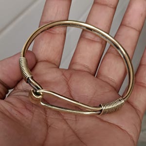 K&ouml;nnte beinhalten: Ein goldfarbenes Metallarmband mit einem einzigartigen Hakenverschluss. Das Armband besteht aus einem d&uuml;nnen, runden Draht, der am Verschluss miteinander verdreht ist.