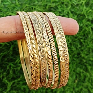 Könnte beinhalten: Satz von sechs goldfarbenen Metallarmbändern mit aufwendigen Prägemustern. Die Armbänder sind übereinander gestapelt und zeigen die verschiedenen Muster.