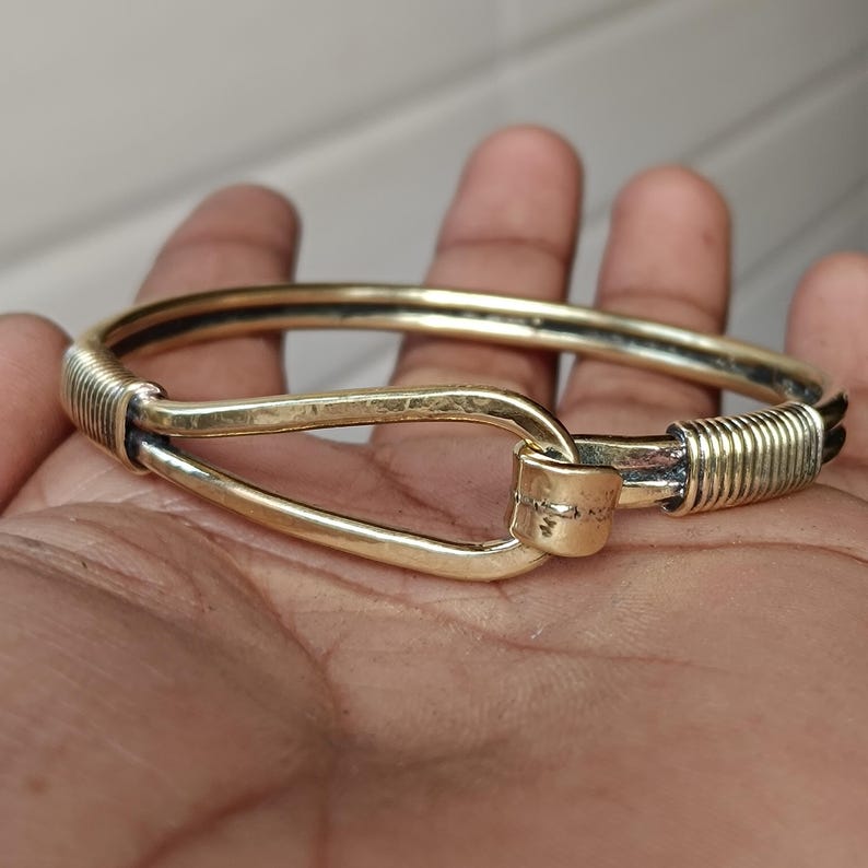 K&ouml;nnte beinhalten: Ein goldfarbenes Armreif-Armband mit einem einzigartigen Verschlussdesign. Das Armband ist aus dickem, strukturiertem Metall gefertigt und hat einen schlichten, eleganten Stil.