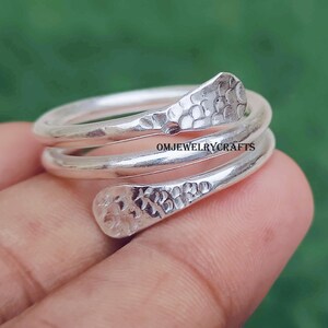 Peut inclure: Une bague en argent enroulée avec une texture martelée aux extrémités. La bague s'enroule autour du doigt, les extrémités se chevauchant. Le texte "OMJEWELRYCRAFTS" est visible sur la bague.