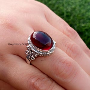 以下が含まれることがあります： 大きな楕円形の深い赤色の宝石をあしらったスターリングシルバーのリング。宝石は、ねじれたロープのデザインと装飾的な装飾が施された、詳細で装飾的なベゼルにセットされています。リングは指に装着されています。