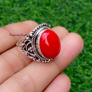 May include: A silver ring with an oval, bright red stone. The ring has an ornate, detailed design around the stone and band. The text "Omjewelry Crafts" is visible on the ring.