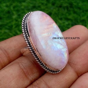 Può includere: Anello in argento con una grande pietra di luna ovale. La pietra di luna mostra un colore rosa pallido e bianco con riflessi iridescenti. L'anello ha una fascia in argento e un design a corda intrecciata attorno alla pietra. Il testo "OMJEWELRYCRAFTS" è visibile.