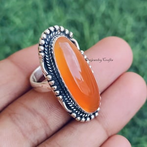 May include: A silver ring featuring an oval, orange gemstone. The gemstone is set in a detailed, ornate silver setting with a beaded border. The ring is held in a hand against a blurred green background. The text "Omjewelry Crafts" is visible.