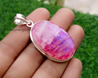 Op de afbeelding: Een zilveren hanger met een ovale, iriserende roze en paarse edelsteen. De edelsteen is gezet in een zilveren bezel en heeft een beugel voor een ketting. De hanger wordt in een hand gehouden tegen een vage groene achtergrond.