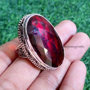 May include: A silver-coloured ring with a large, oval, deep red gemstone. The ring has an ornate setting with detailed filigree and decorative accents. The faceted gemstone is the focal point, catching the light and showcasing its rich colour.