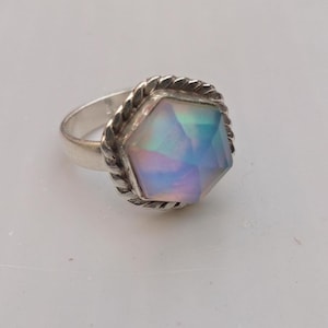 May include: A silver ring with a large, iridescent, faceted gemstone. The gemstone has a hexagonal shape and displays a range of colors, including blue, purple, and pink. The ring's band is simple, while the gemstone is framed by a twisted rope design.