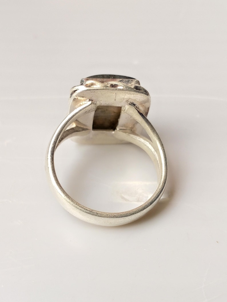 Op de afbeelding: Een zilveren ring met een rechthoekige, donkergekleurde steen. De ring heeft een eenvoudige band en een decoratieve zetting rond de steen. De ring wordt getoond tegen een witte achtergrond.