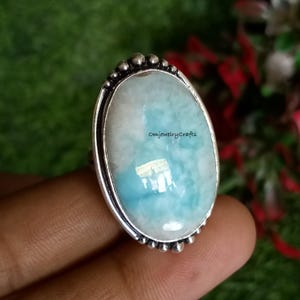 May include: Oval-shaped ring with a light blue and white gemstone set in a silver band. The band is decorated with small beads around the edge. The gemstone has a cloudy, iridescent appearance.