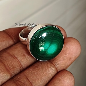 May include: A silver ring with a large, round malachite stone. The malachite is a deep green with swirling patterns. The ring is set in a simple, polished silver band. The text "OmjewelryCrafts" is visible.