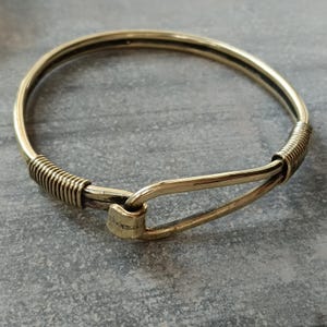 K&ouml;nnte beinhalten: Ein goldfarbenes Armreif-Armband mit einem einzigartigen Verschlussdesign. Das Armband besteht aus einem dicken, runden Draht und hat an einer Seite einen gewickelten Abschnitt.