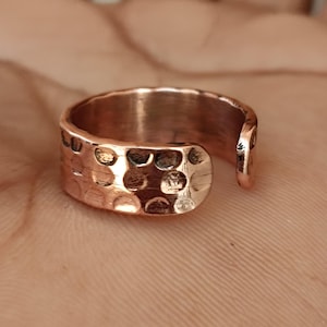 Op de afbeelding: Een koperkleurige verstelbare ring met een gehamerde textuur. De ring heeft een afgeronde band en een open ontwerp, waardoor de maat kan worden aangepast. De gehamerde textuur creëert een unieke, handgemaakte uitstraling. De ring wordt getoond op een huidkleurige achtergrond.