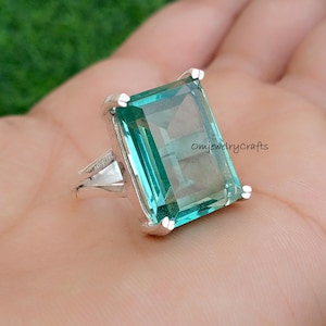 Può includere: Un anello d'argento con una grande pietra preziosa verde smeraldo incastonata in una montatura quadrata, a bezel. L'anello è su una mano, e il testo "OmjewelryCrafts" è visibile sullo sfondo.