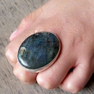 May include: A silver ring featuring a large oval labradorite gemstone. The stone displays a dark blue and green hue with iridescent flashes of light.
