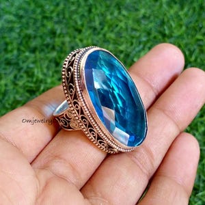 Könnte beinhalten: Ein Ring mit einem großen, ovalförmigen blauen Edelstein. Der Ring hat eine aufwendige, filigrane Fassung und ein silberfarbenes Band. Der Hintergrund ist grün.