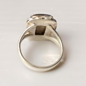 Op de afbeelding: Een zilveren ring met een rechthoekige, donkergekleurde steen. De ring heeft een eenvoudige band en een decoratieve zetting rond de steen. De ring wordt getoond tegen een witte achtergrond.